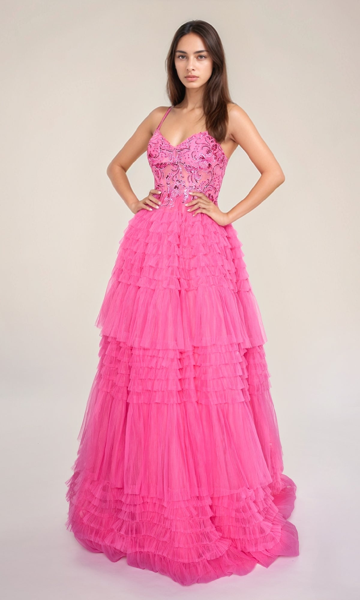 A woman stands with hands on hips, modeling the Nina Canacci 1582 long formal dress in bright pink, featuring an A-line ruffle tiered skirt and embellished fitted bodice, against a plain light background.